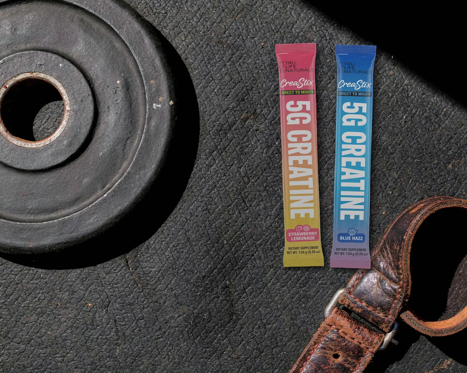Strawberry Lemonade and Blue Razz CreaStix sticks on a rubber gym floor beside a weight plate and a leather lifting belt