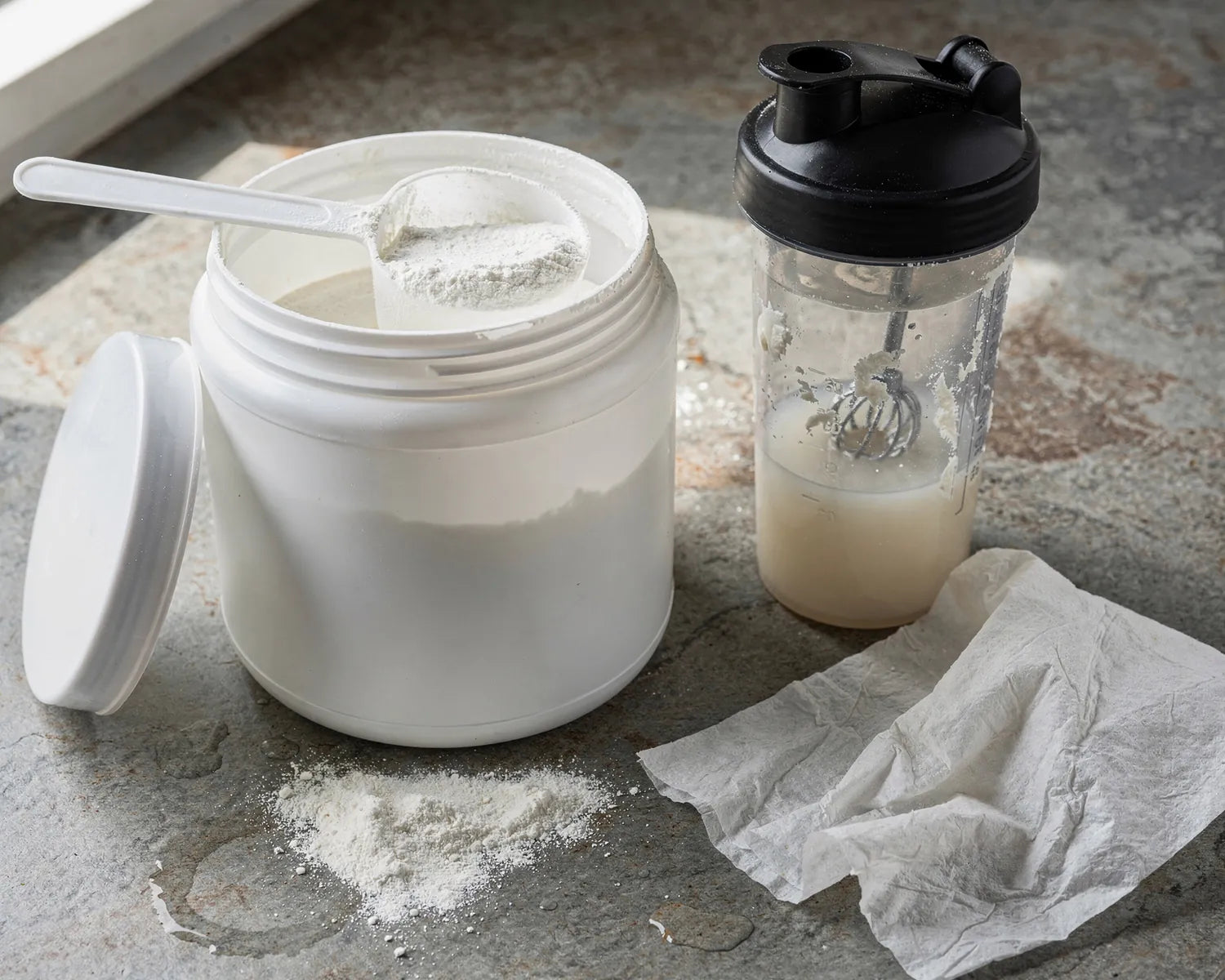 A tub of creatine powder with a heaped scoop, a shaker bottle with clumps, and spilled powder
